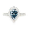 A picture of a blue pear cut moissanite engagement ring with a diamond halo and a pavé band.